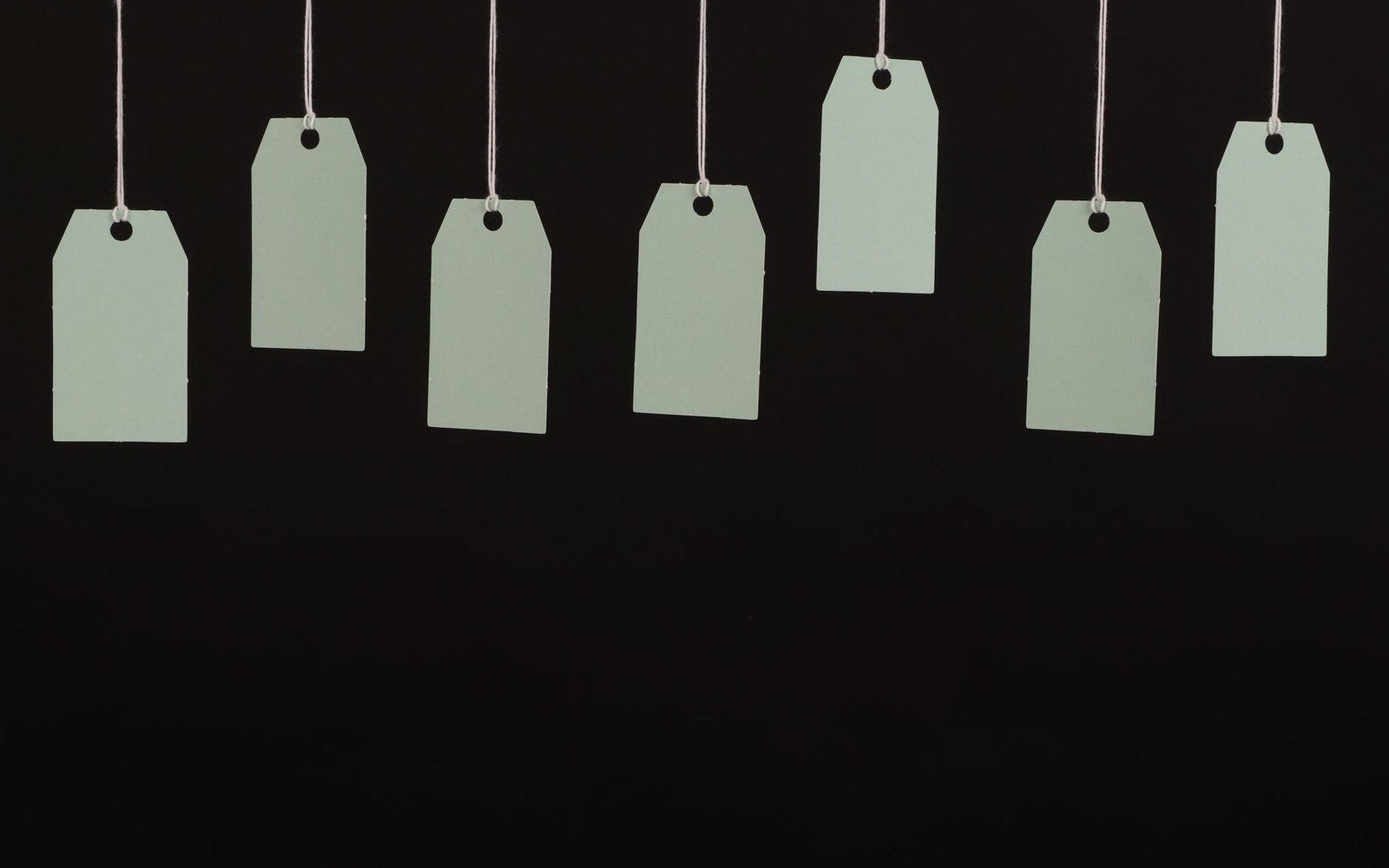 Seven blank pale green price tags hanging on strings against a black background